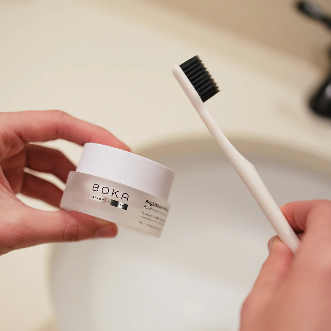 Hand holding a black toothbrush and a jar of Boka toothpaste in front of a bathroom sink.