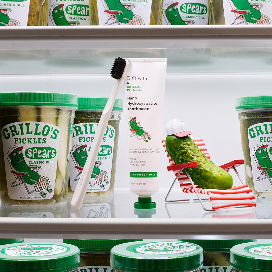 A pickle relaxes on a chair beside toothpaste and a toothbrush, inside a fridge with jars of pickles.