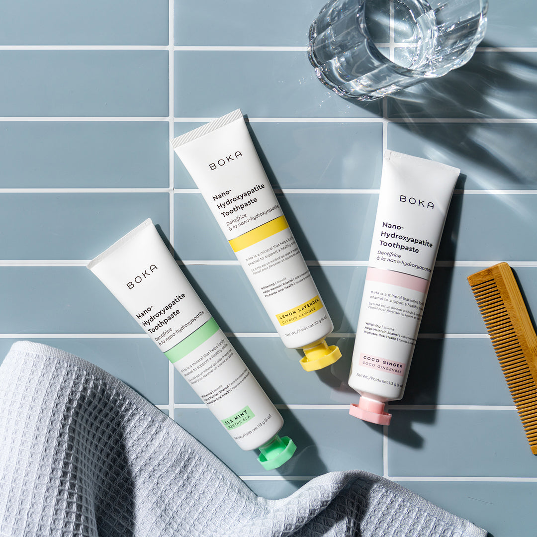 Three Boka toothpaste tubes, a glass of water, a yellow comb, and a towel on blue tiles.