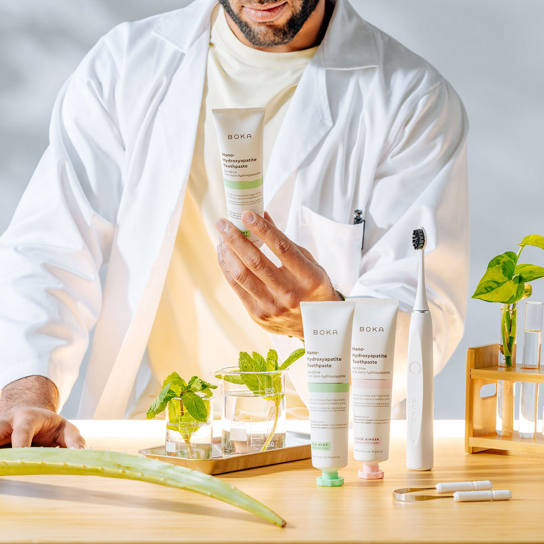 A person in a lab coat displays Boka toothpaste with plants, a toothbrush, and lab glassware on a table.
