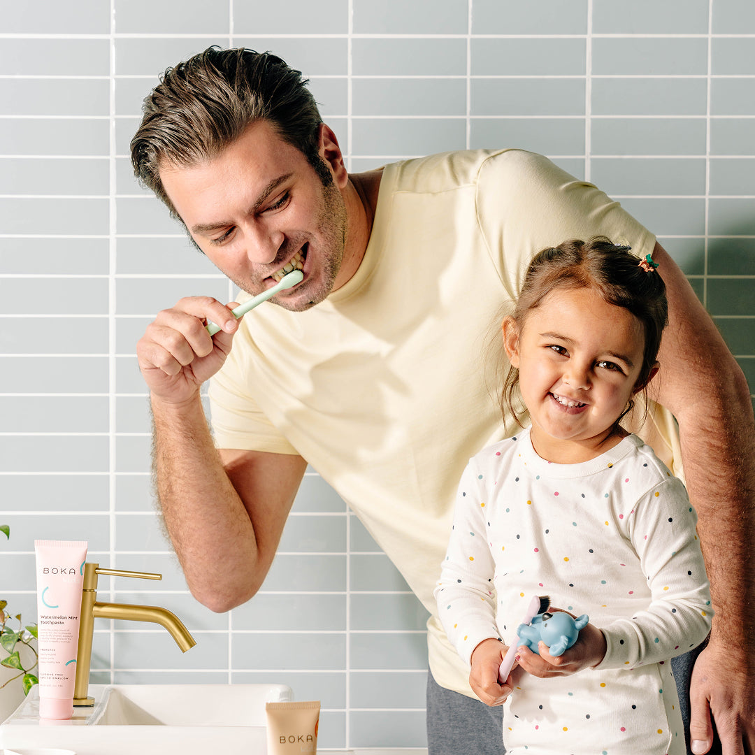 Smiling man brushes teeth at sink while young girl holding a toy smiles at the camera.