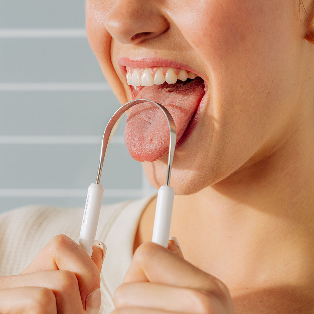 A person using a metal tongue scraper to clean their tongue, smiling with mouth open.