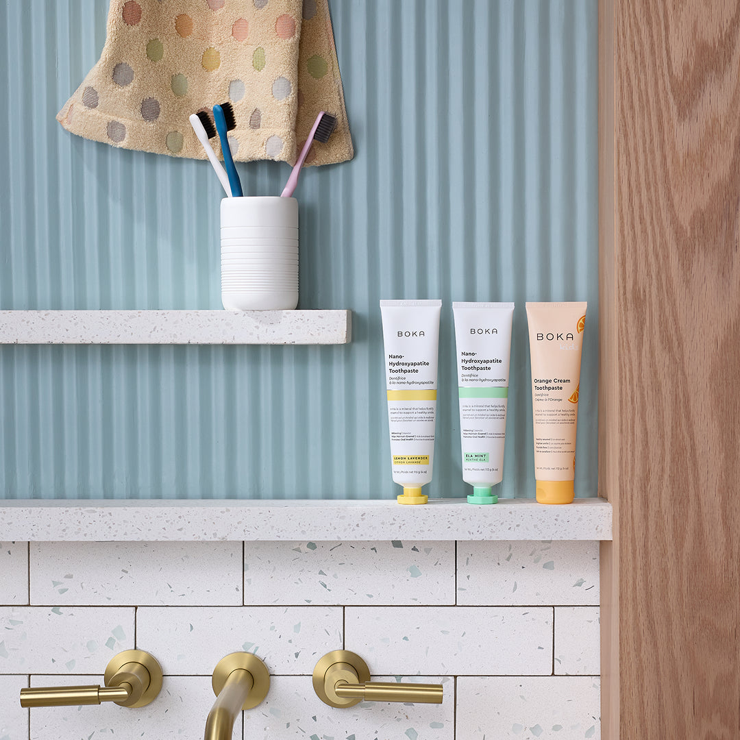 Three tubes of Boka toothpaste and a cup with toothbrushes on a bathroom shelf above a sink.