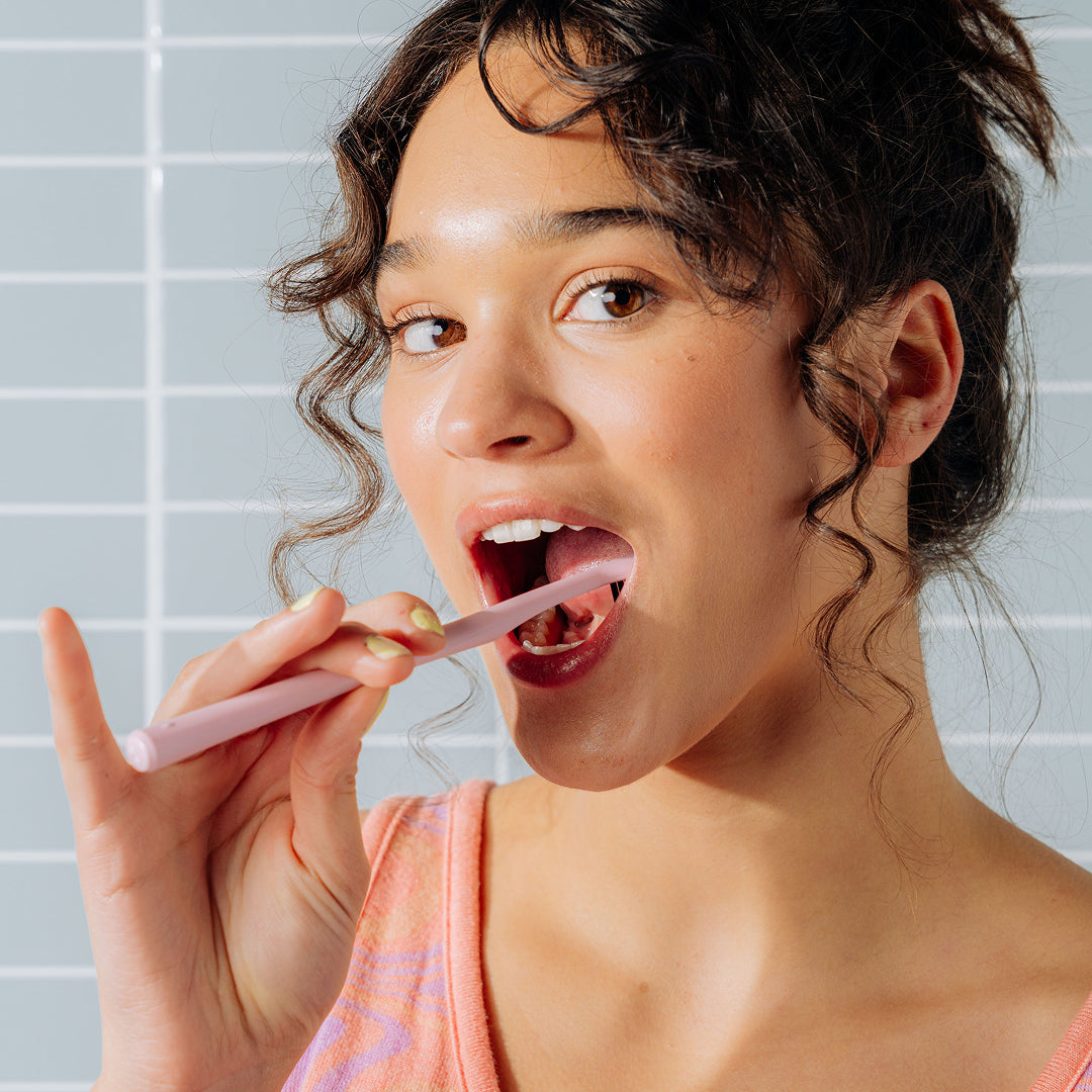 Woman brushing her teeth with a pink toothbrush in front of a light blue tiled wall.