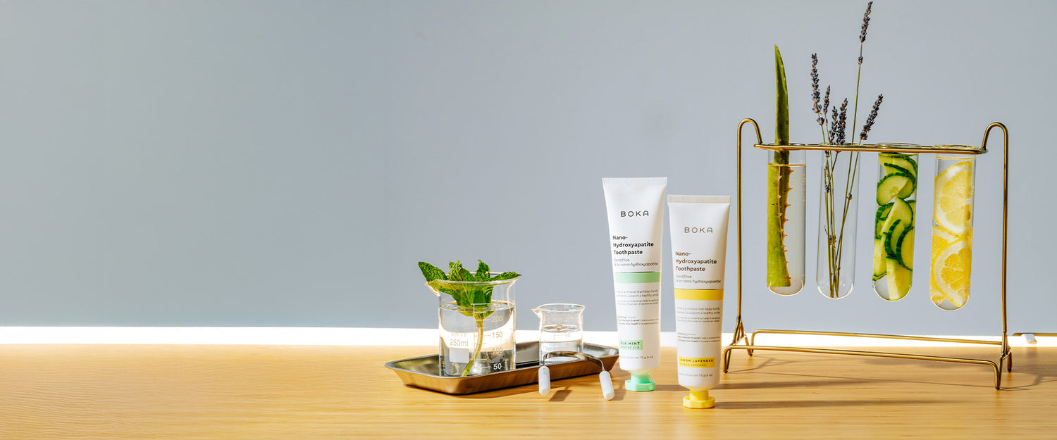 Two tubes of Boka toothpaste next to glassware with mint, cucumber, lemon, and lavender stems.