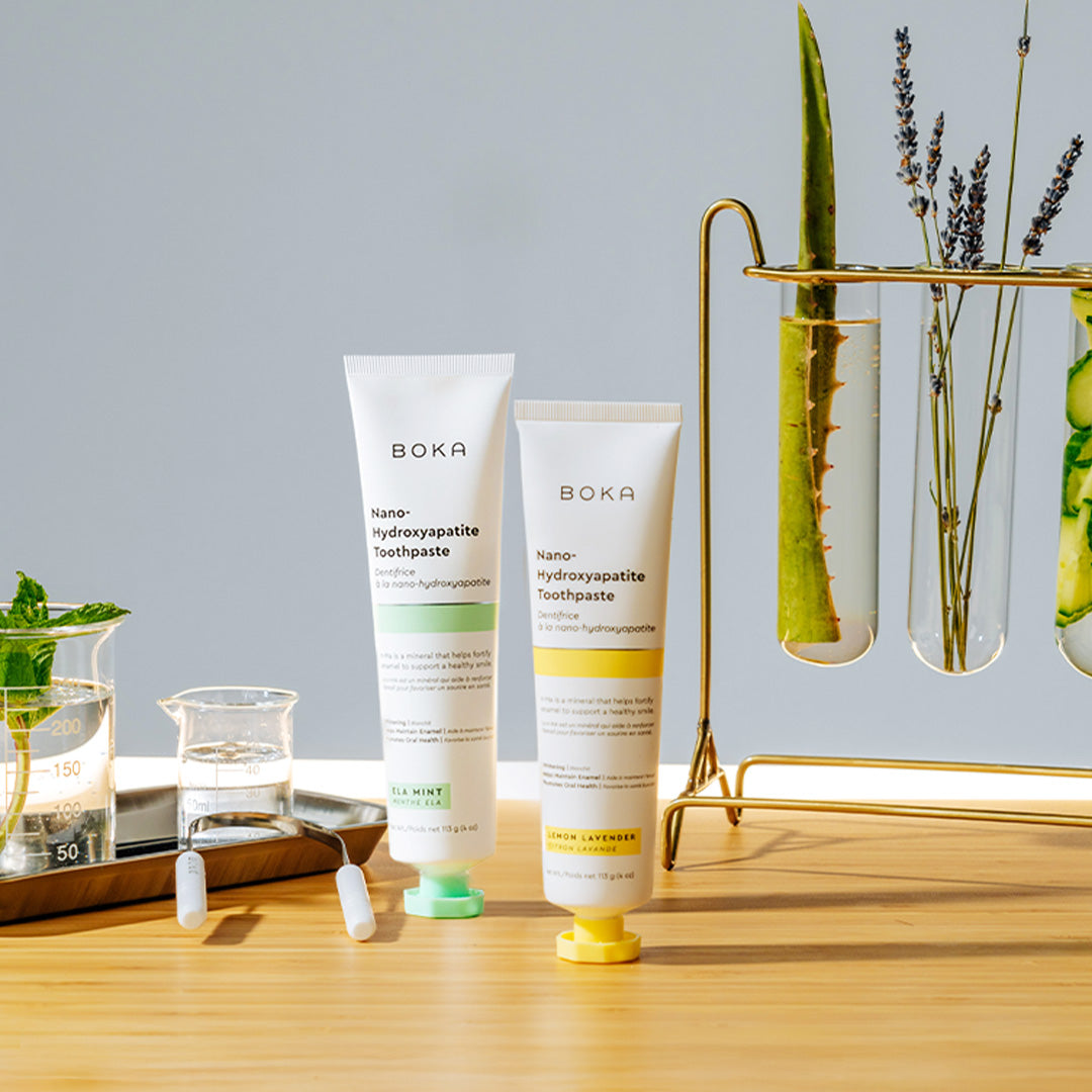 Two Boka toothpaste tubes with mint, herbs, and lab glassware in the background on a light surface.