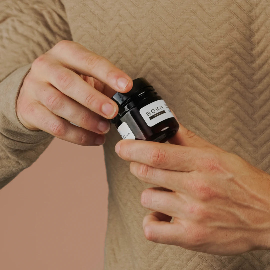 A person in a beige top holds and opens a small jar labeled BOKA against a neutral background.