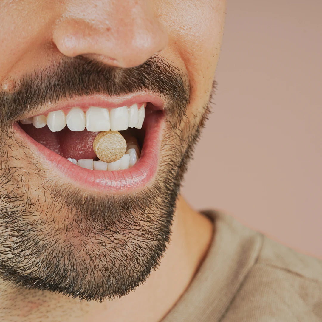 A man holds a round tablet between his teeth, smiling with his mouth slightly open.