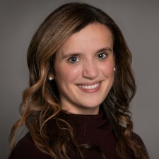 Woman with long wavy brown hair smiling, wearing a dark top, posed in front of a gray background.