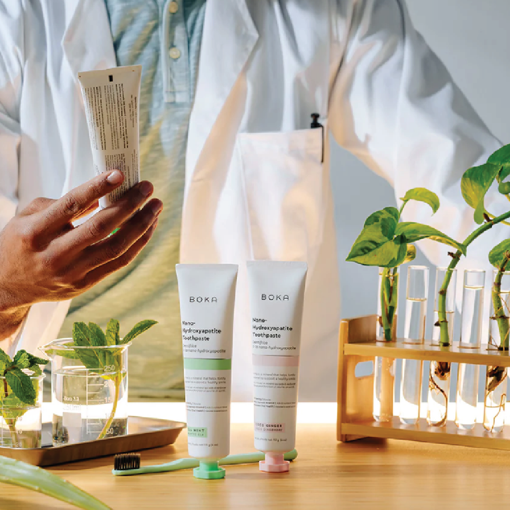 Person in lab coat holding a tube beside toothpaste, plants, glassware, and test tubes on a table.