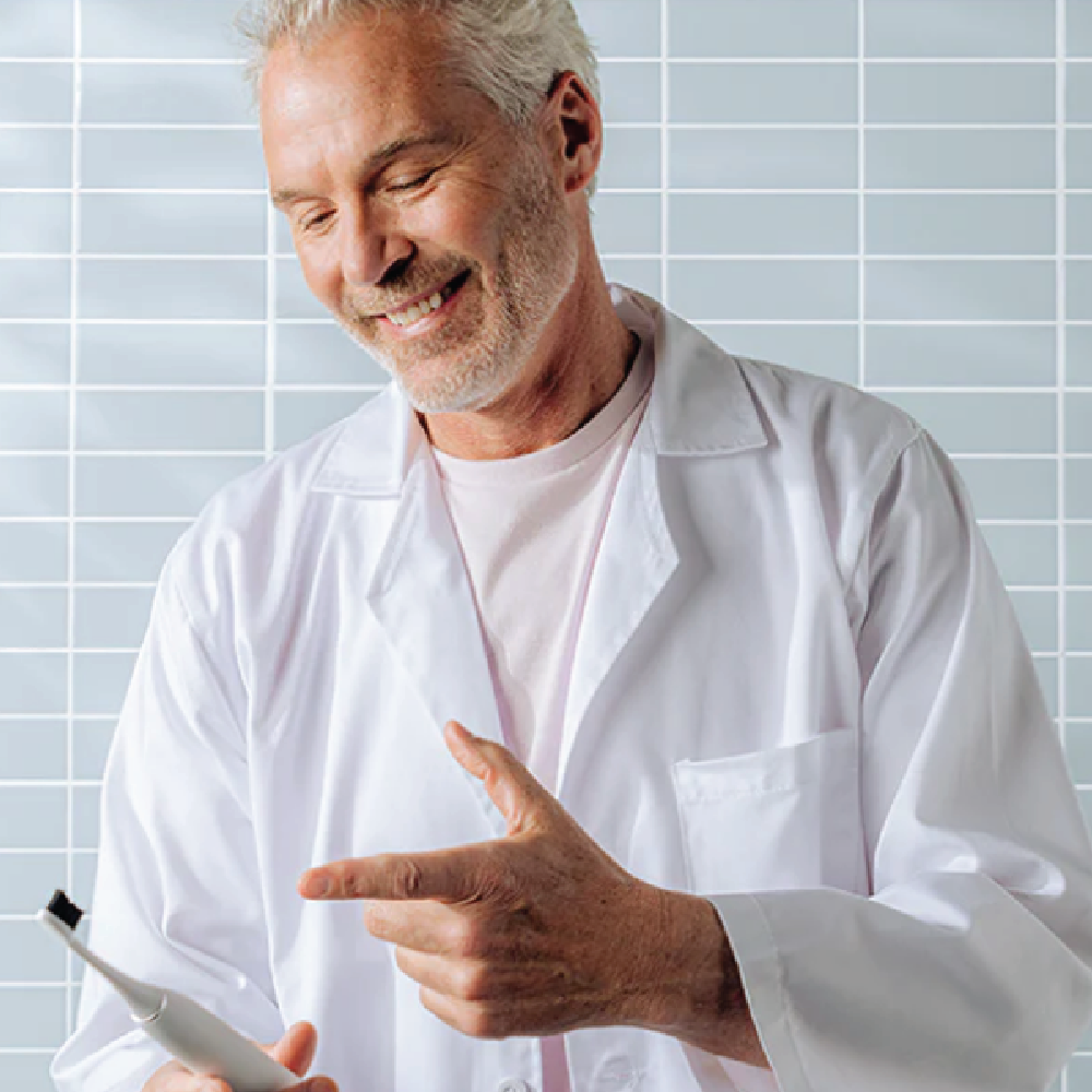 Smiling man in a white lab coat pointing at an electric toothbrush against a tiled wall.